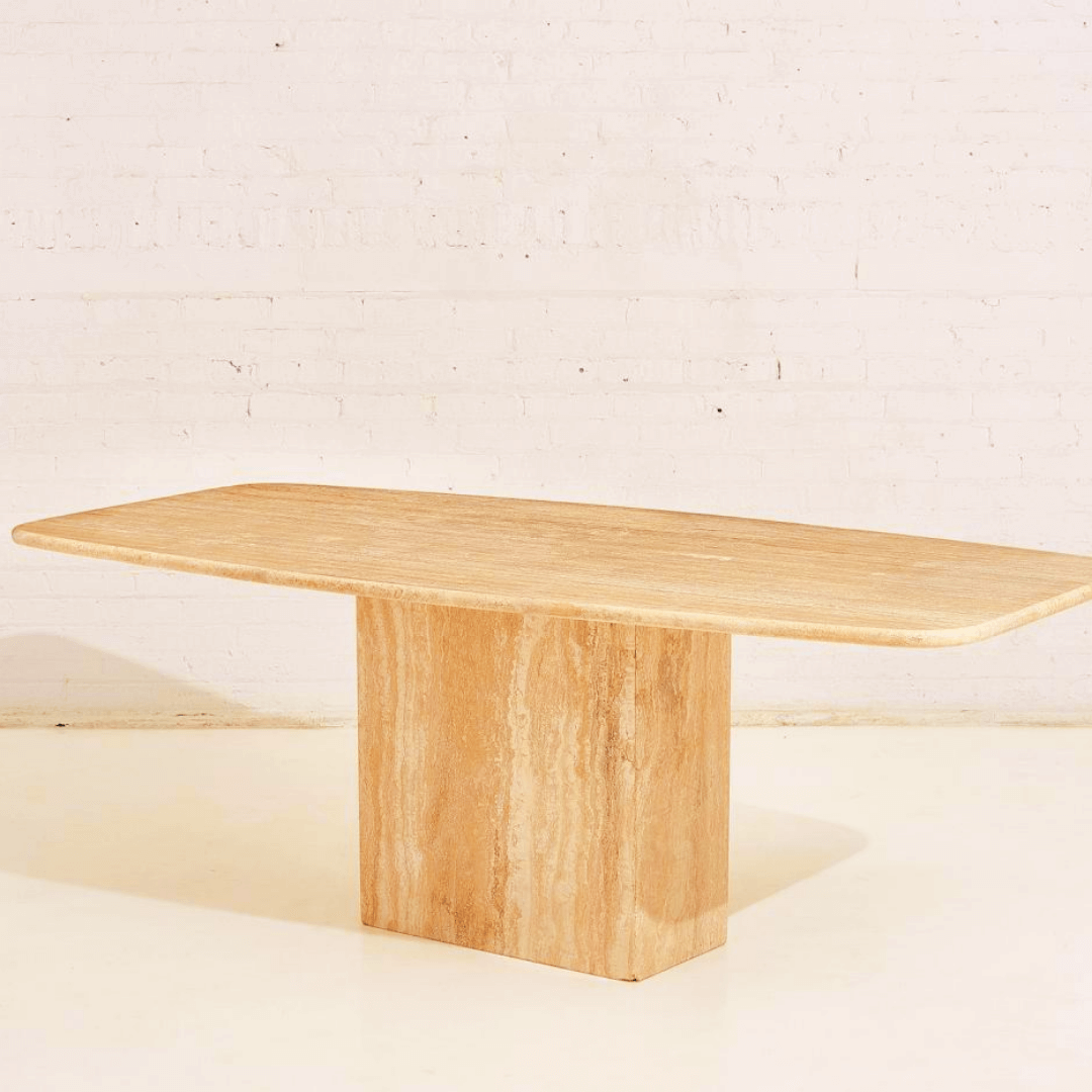 A minimalist rectangular wooden table with a slab top and block base stands against a light brick wall and floor, reminiscent of Travertables Serenity Travertine Dining Table. Its elegant form mirrors the premium travertines beauty, casting a faint shadow to the left.