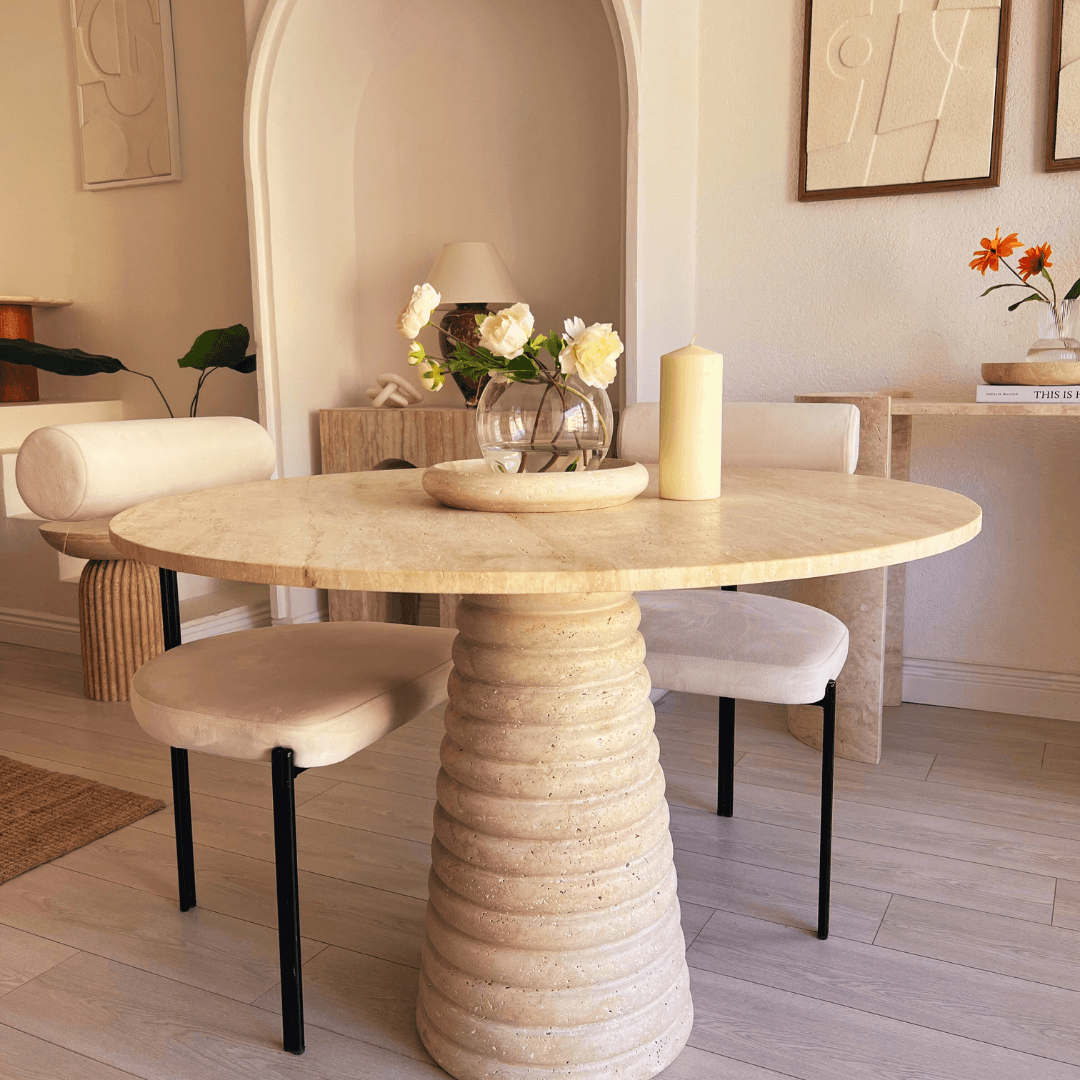 Set in a modern, minimalist room, The Harmony Round Travertine Dining Table by Travertable features a stone-textured surface and sculptural base. Adorned with white flowers, a candle, two curved cushioned chairs, and surrounded by neutral decor and abstract art for customizable style.