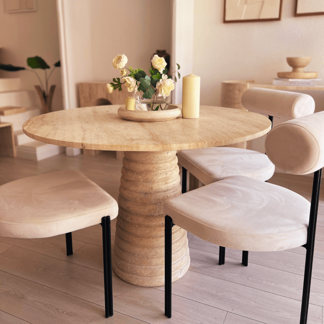 The Harmony Round Travertine Dining Table by Travertable, with its textured base, is encircled by four beige cushioned chairs. A vase of yellow and white flowers and two candles embellish the table, set against a neutral-toned interior with framed art and a small sculpture.