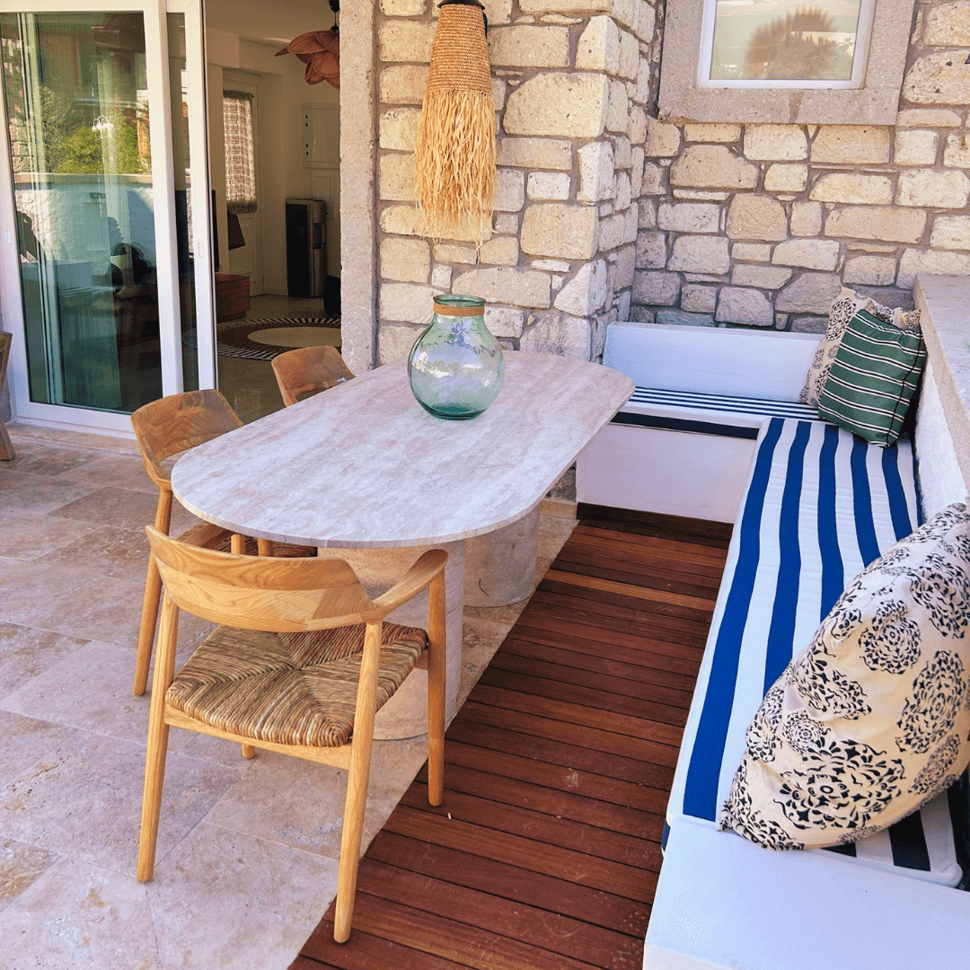 The outdoor patio, framed by a stone wall, features an Arcadia Dining Table by Travertable, surrounded by wicker chairs and a cushioned blue-and-white striped bench with patterned pillows. A large glass vase adorns the travertine surface as natural light filters through the glass doors.