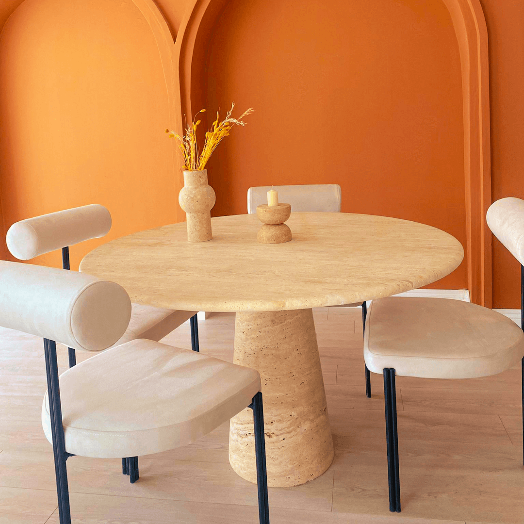 The Elysian Travertine Round Dining Table by Travertable holds a vase of yellow flowers and a small candle holder, surrounded by four beige cushioned chairs. A stylish orange wall with arched recesses enhances the contemporary look.