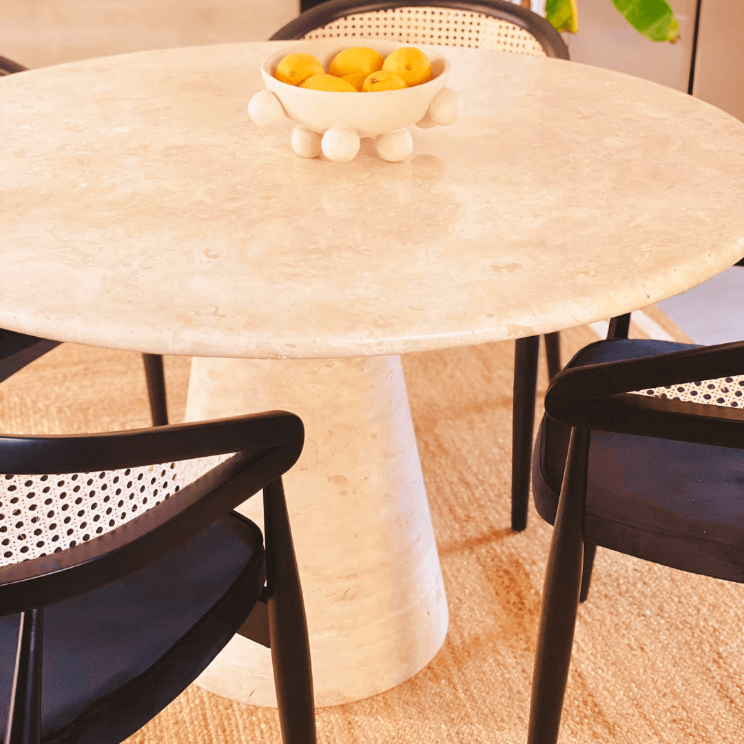 An Elysian Travertine Round Dining Table by Travertable with a pedestal base features a decorative bowl of lemons. Four black chairs with woven cane backs complement the modern setup, all resting on a textured beige rug.