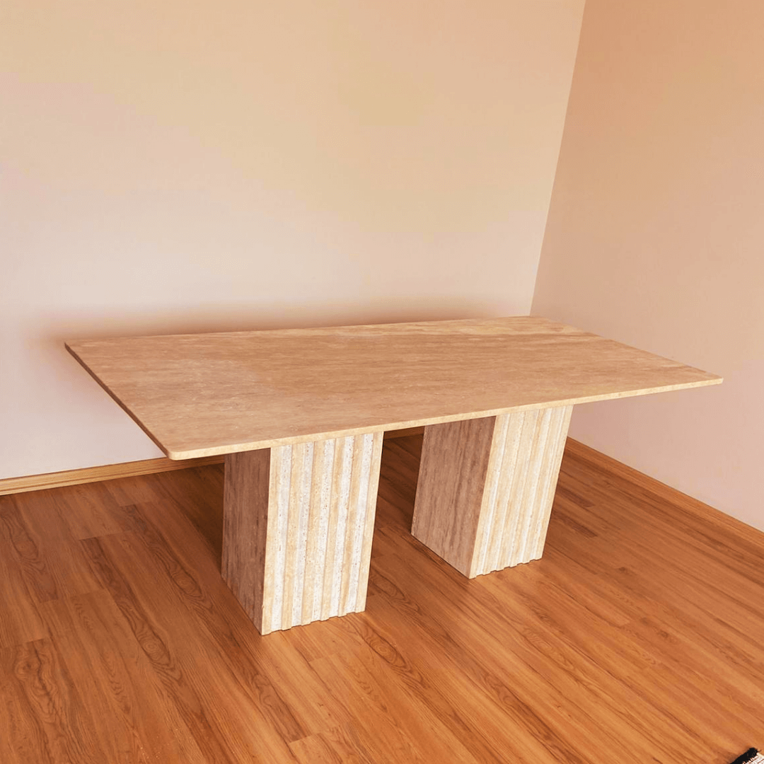 The Ridge Rectangular Travertine Dining Table by Travertable showcases modern sophistication with its stone-like surface, two columnar supports, and fluted legs featuring elegant vertical grooves. It stands on a light wooden floor against a plain wall.