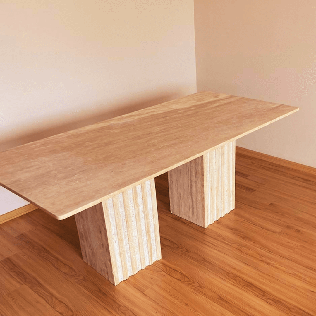 The Ridge Rectangular Travertine Dining Table by Travertable exudes modern sophistication with its sleek top and two wide, fluted pedestal legs, standing gracefully on a hardwood floor against a plain light-colored wall.