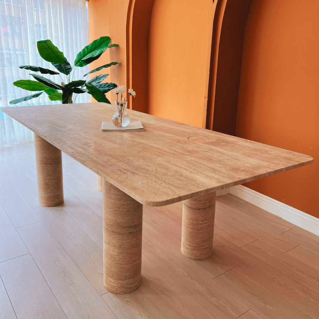 The Columnar Travertine Dining Table by Travertable, set in a warmly lit room with orange walls and white curtains, features contemporary design with cylindrical legs. A nearby green plant adds a touch of nature, while a small vase and book on the table offer customizable decoration options.
