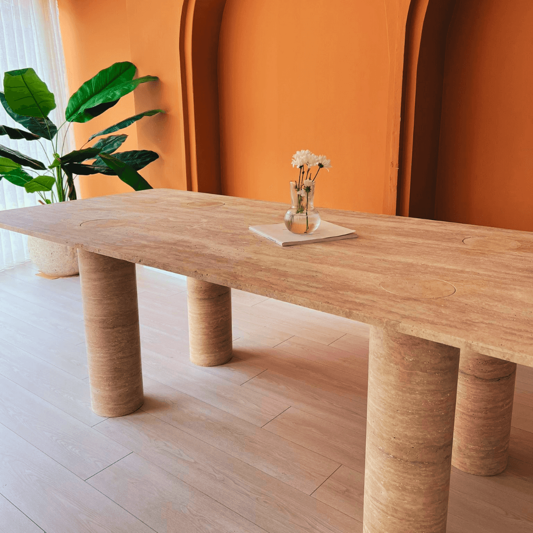 In a modern room, a wooden table with cylindrical legs mirrors the elegance of Travertables The Columnar Travertine Dining Table, topped with a vase of white flowers. Warm orange walls complement the table and large green plant, enhancing the contemporary charm over light wood flooring.