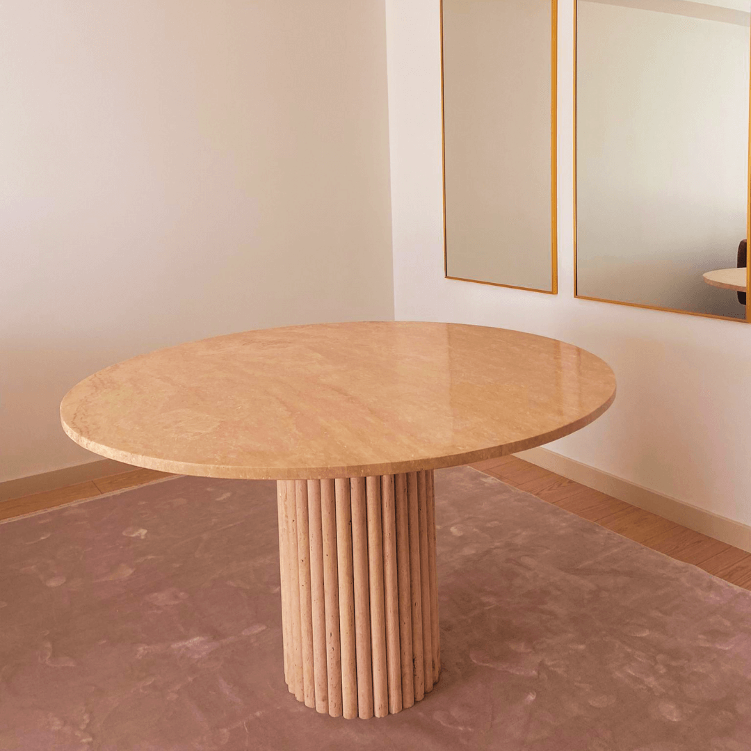 The Classic Round Travertine Dining Table by Travertable, featuring a textured cylindrical base, stands on a light brown rug in this minimalist room. The white walls and large mirror reflect the bespoke piece and the elegant wall decor.
