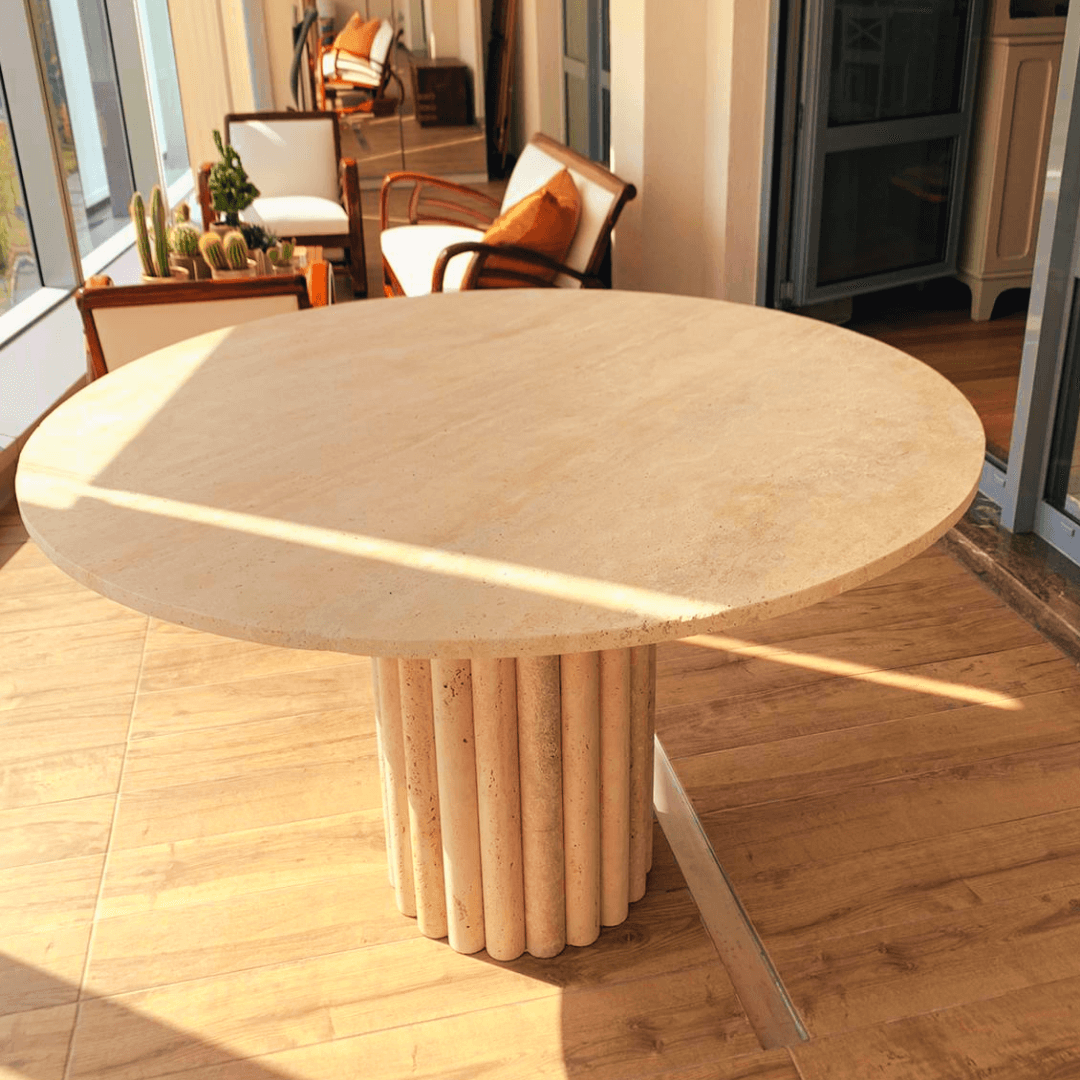 The Classic Round Travertine Dining Table by Travertable, featuring a ribbed cylindrical base, sits on a wooden floor. Sunlight highlights its minimalist design while nearby cushioned chairs and small potted cacti enhance the space near a large window.