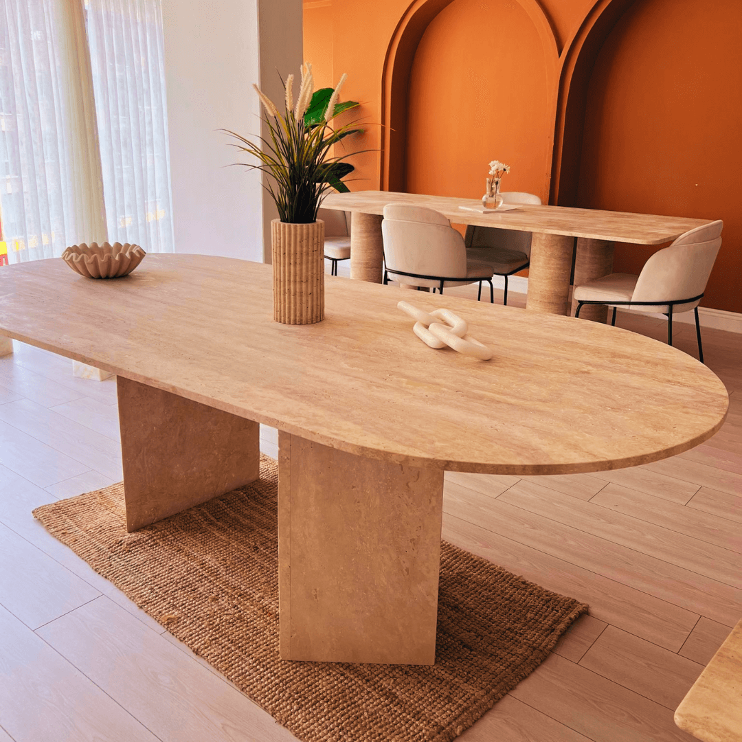 A stylish room showcases Travertables Grand Pillar Travertine Dining and Meeting Table on a woven rug, adorned with a plant-filled vase and decor. Beige chairs encircle another table against a burned orange arched wall. Sheer curtains let natural light in, enhancing the modern aesthetic.