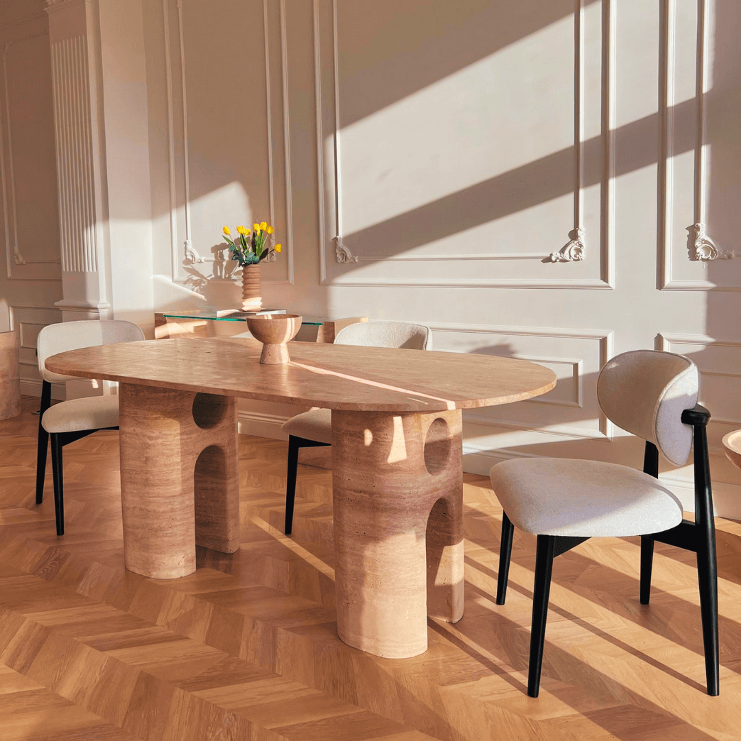 The dining room showcases a Travertables Travertine Majesty Ellipse Table with three gray upholstered chairs on a parquet floor. A vase of yellow flowers adorns the table, while sunlight highlights the elegant white paneled walls.