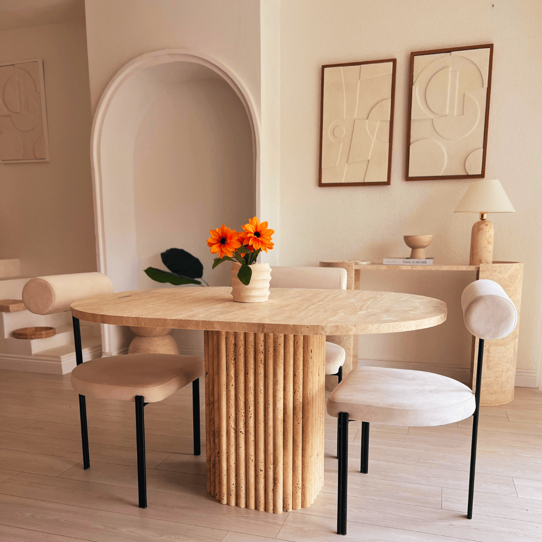 A modern dining room features the Harmony Oval Travertine Dining Table by Travertable, surrounded by minimalist chairs. Orange flowers in a white vase elevate the tables elegance. Neutral-toned wall art and a small side table with a lamp complete the cozy space.