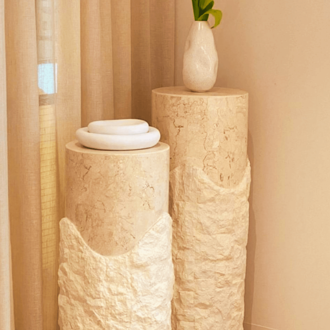 Two Rustic Charm Travertine Pedestals by Travertable stand against a curtain backdrop. A white vase with green leaves is on the taller one, while stacked white rings adorn the shorter, crafting a minimalist and modern display of sculptural columns.