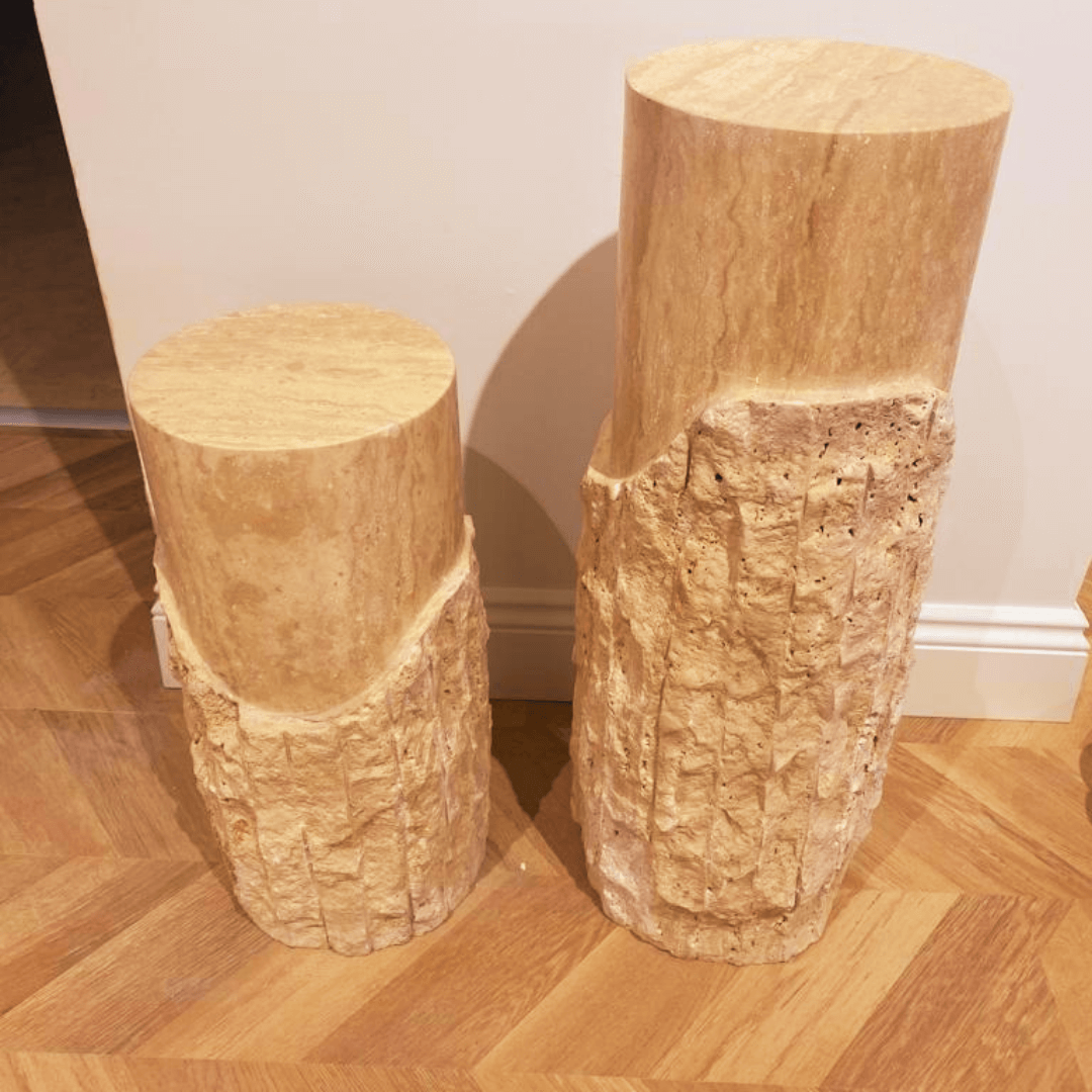 On a wooden herringbone-patterned floor, two Travertable Rustic Charm Travertine Pedestals are displayed. Made of textured, light stone with smooth tops and uneven sides, one sculptural column stands taller than the other, adding visual intrigue.