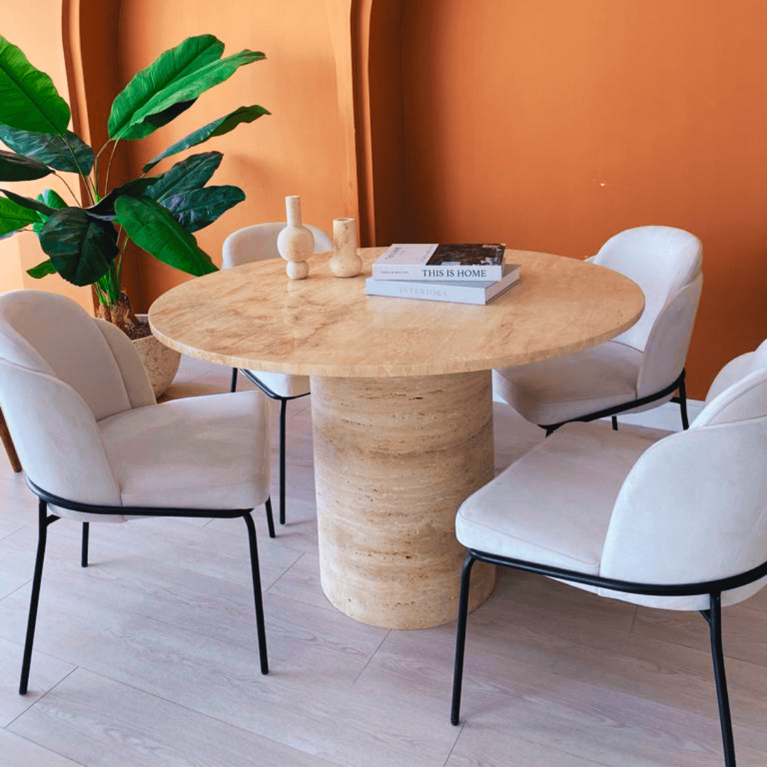 The Elysian Travertine Round Dining Table by Travertable features a beige stone surface surrounded by four white cushioned chairs with black metal legs. Two small vases and books sit on the customizable table, while a large potted plant and warm terracotta walls complete the setting.
