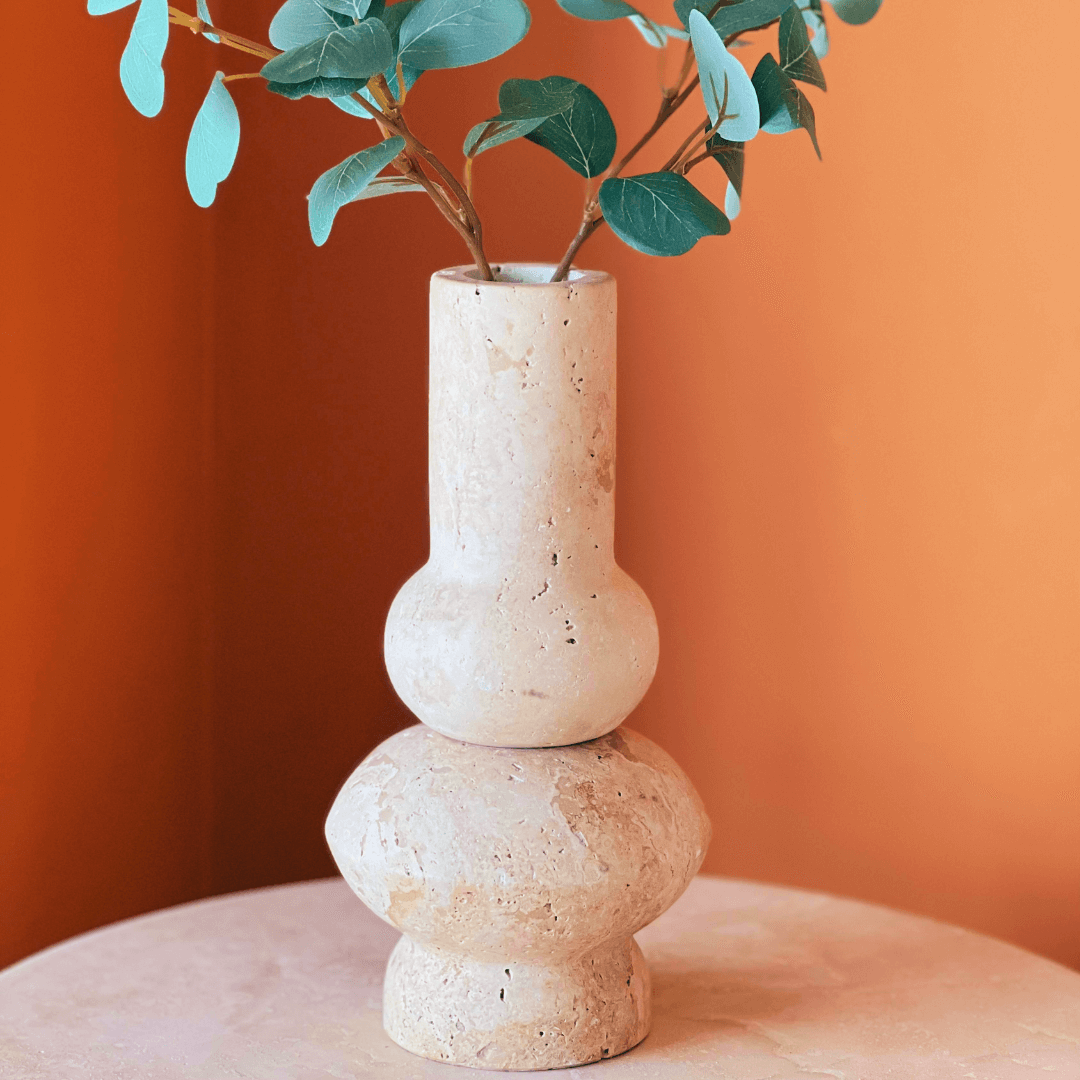 A Minimalist Beige Travertine Vase by Travertable, featuring a unique, curvy shape and textured ceramic finish, holds green eucalyptus stems on a matching table against a warm orange background.