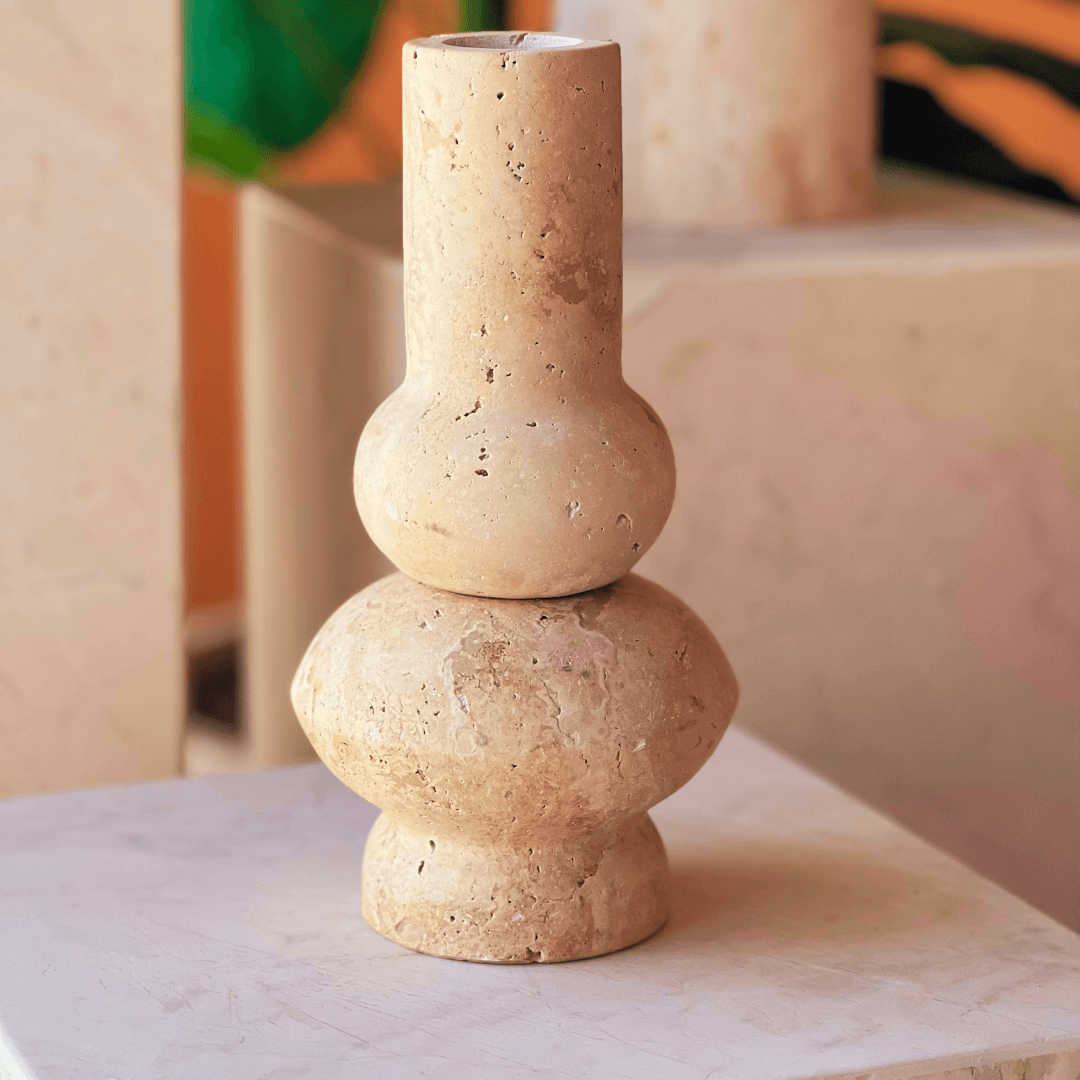 The Minimalist Beige Travertine Vase by Travertable features a cylindrical top and rounded bottom with natural beige tones and textured surface, accentuated by soft lighting against a blurred green and orange backdrop.