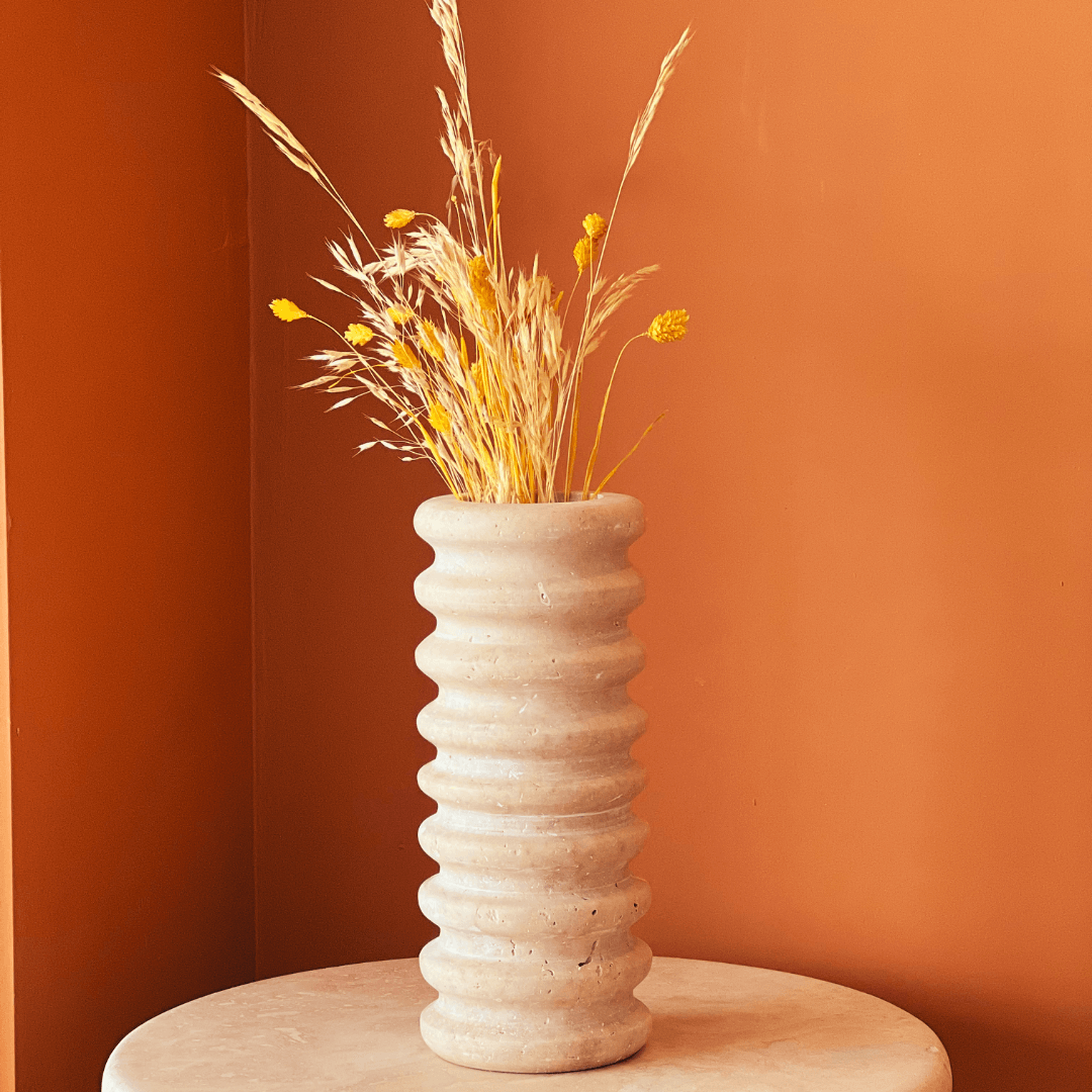 The Natural Beauty Oval Travertine Vase by Travertable, filled with dried yellow and brown grasses and flowers, sits on a circular beige table against a warm orange wall.