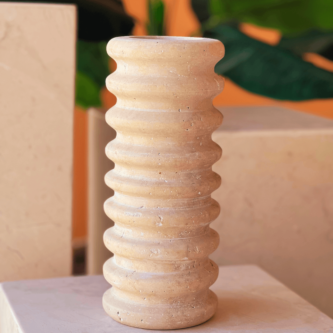 The Natural Beauty Oval Travertine Vase by Travertable features a wavy-textured concrete design in beige, displayed on a light surface. Backdrop includes abstract stone shapes and green foliage.