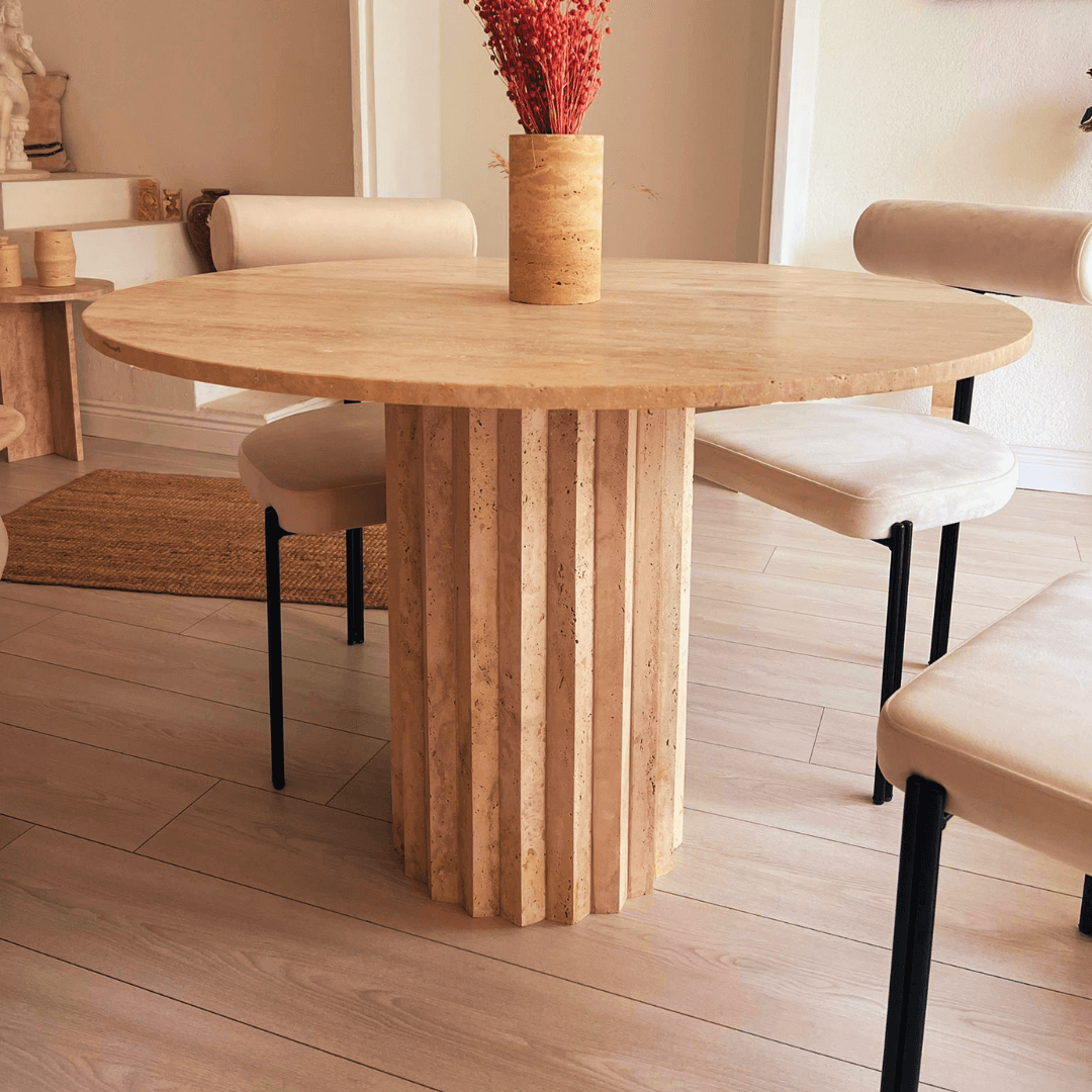 The Fluted Round Travertine Dining Table by Travertable is surrounded by four cream-colored chairs, with a tall vase of red dried flowers at its center. The light wood floor and neutral decor in the room beautifully complement the tables timeless elegance.