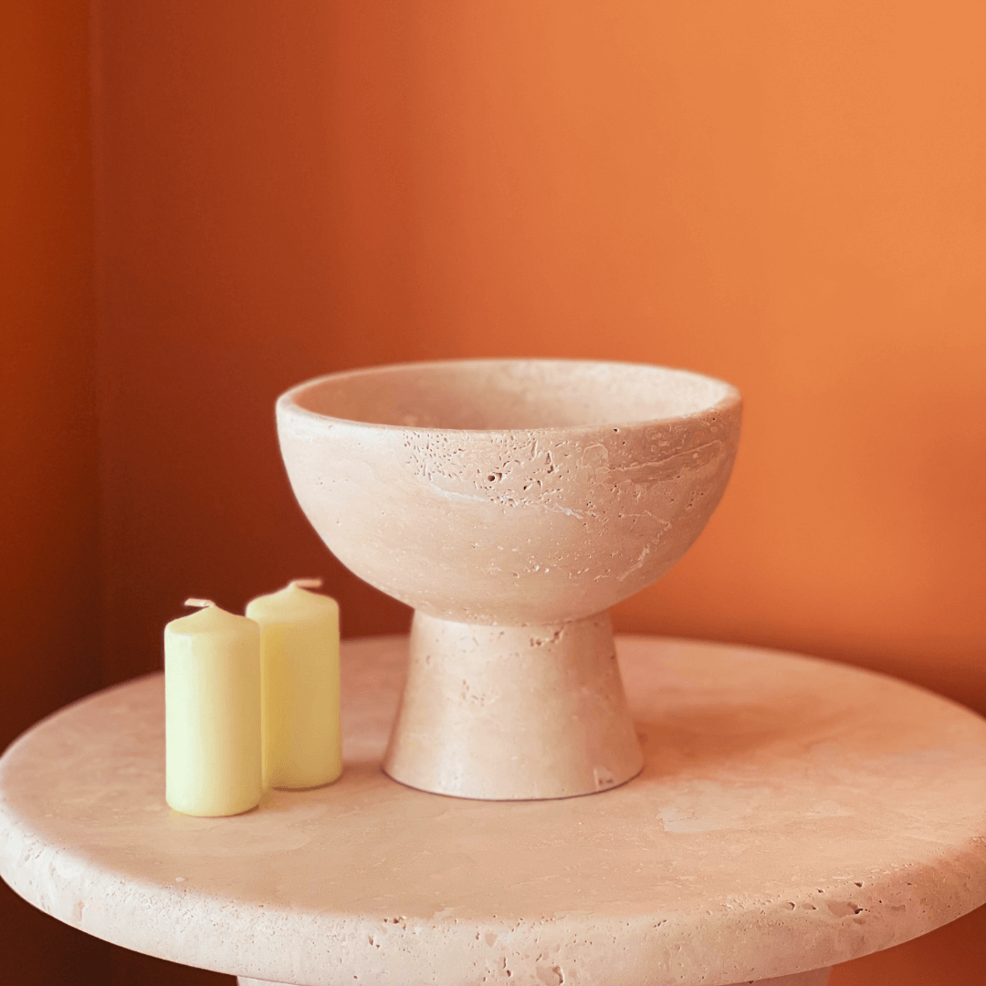 The Sleek Modern Travertine Marble Bowl by Travertable sits on a matching light beige circular table, accompanied by two unlit, pale yellow pillar candles, all set against a warm orange wall.