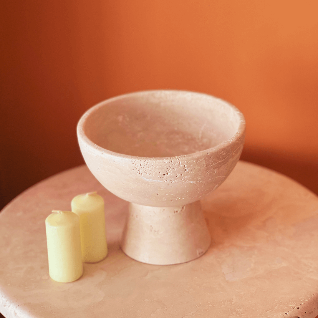 A Sleek Modern Travertine Marble Bowl from Travertable, featuring a wide rim and narrow base, is displayed on a matching marble surface. Two light yellow candles are placed beside it, set against a warm orange background.
