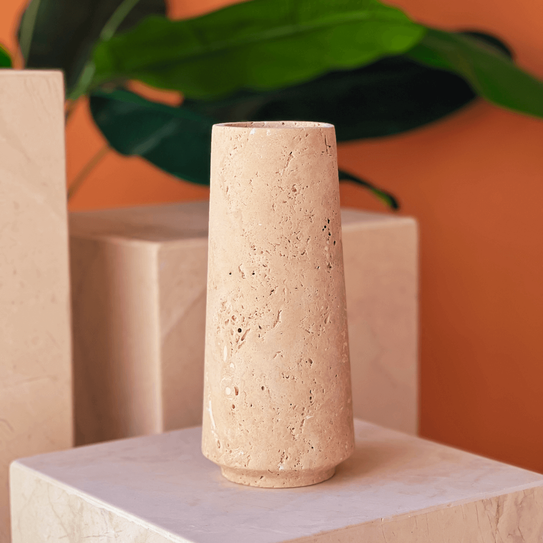 The Classic Beige Travertine Vase by Travertable sits tall on a marble platform, against an orange background with large green leaves.