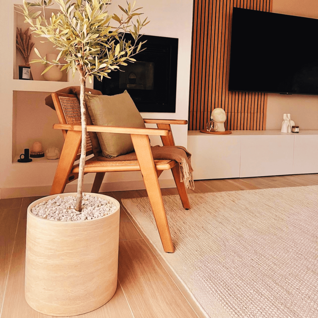 A modern living room includes a wooden armchair with green and brown cushions next to a large potted plant, complementing the Golden Sunrise Travertine Vase by Travertable. Wooden paneling and a white TV console with a flat-screen TV accentuate the scene, while a textured rug adorns the floor.