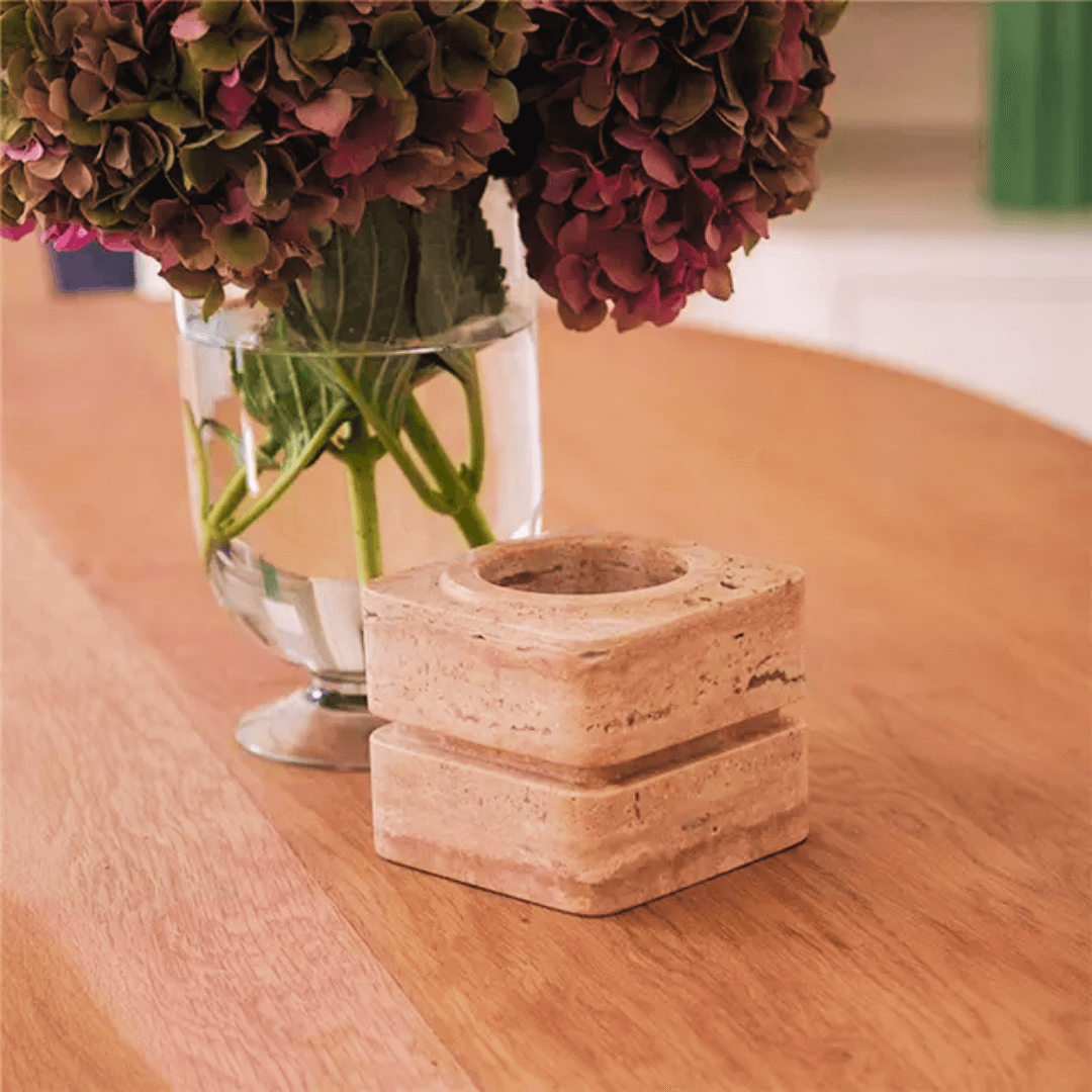 A Vintage Charm Travertine Vase by Travertable, filled with pink and purple hydrangeas, sits alongside a square stone candle holder on a wooden table. The setting offers a warm and cozy ambiance with an emphasis on natural materials.