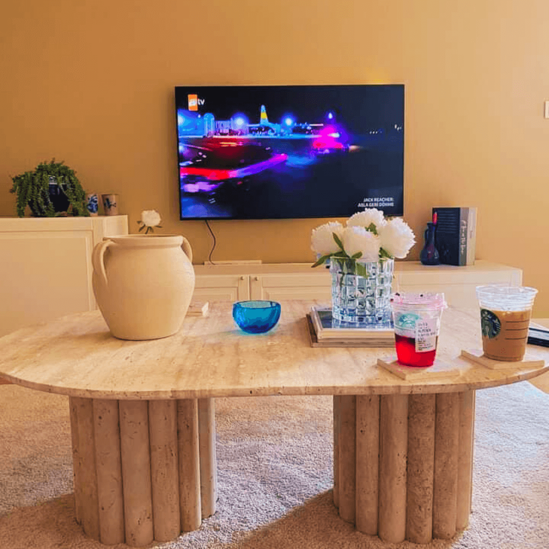 A cozy living room features the Travertable Handcrafted Italian Travertine Coffee Table, known for its architectural presence, adorned with a vase of white flowers, a pitcher, and two drink cups. A flat-screen TV displays a vibrant scene above a cabinet enhanced with a plant and decorative book.
