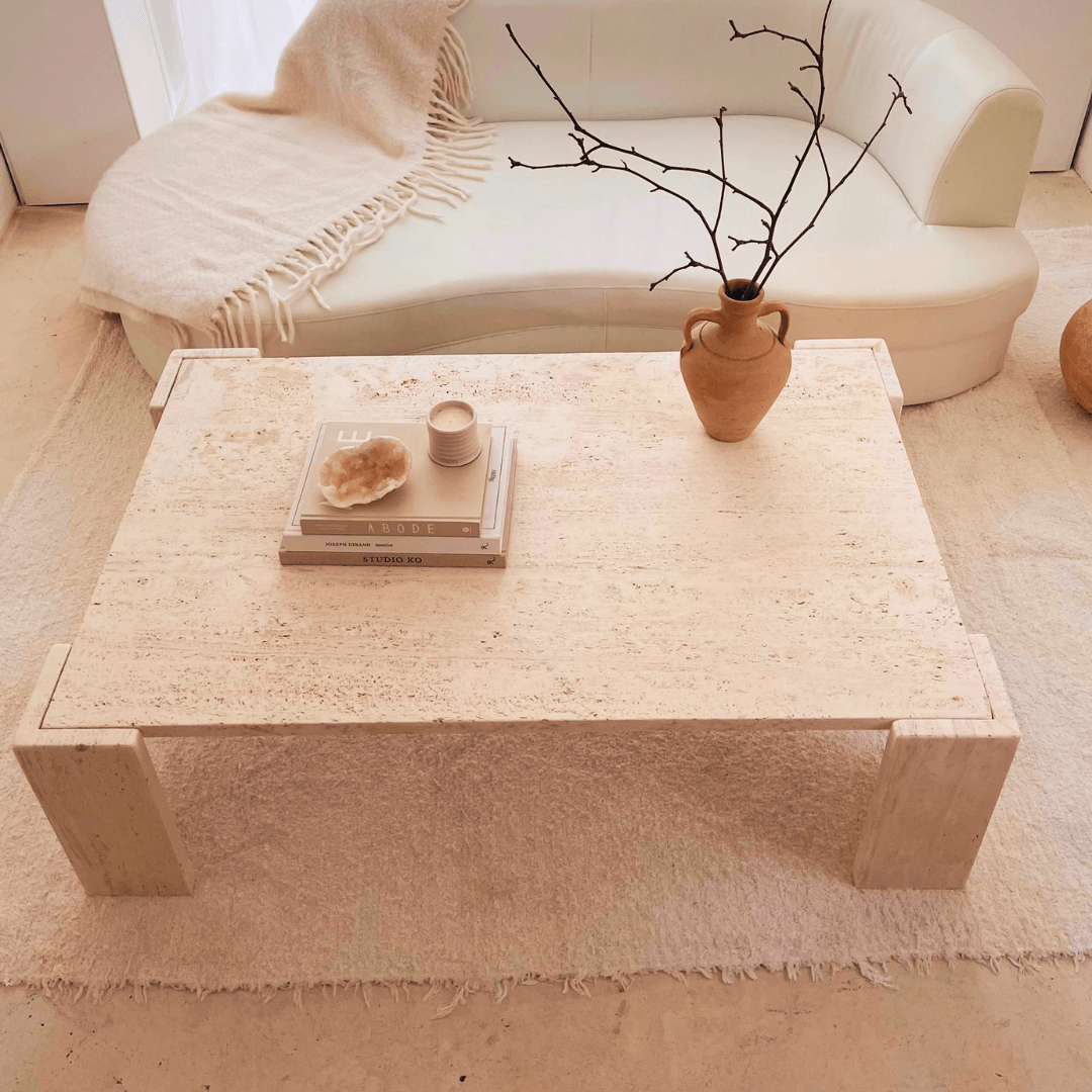 A minimalist living room showcases a white sofa and a Modern Rustic Travertine Coffee Table by Travertable, adorned with a vase of twigs, sculpture, candle, and stacked books. A textured rug and blanket over the sofa highlight the natural beauty of this expertly crafted cozy setting.