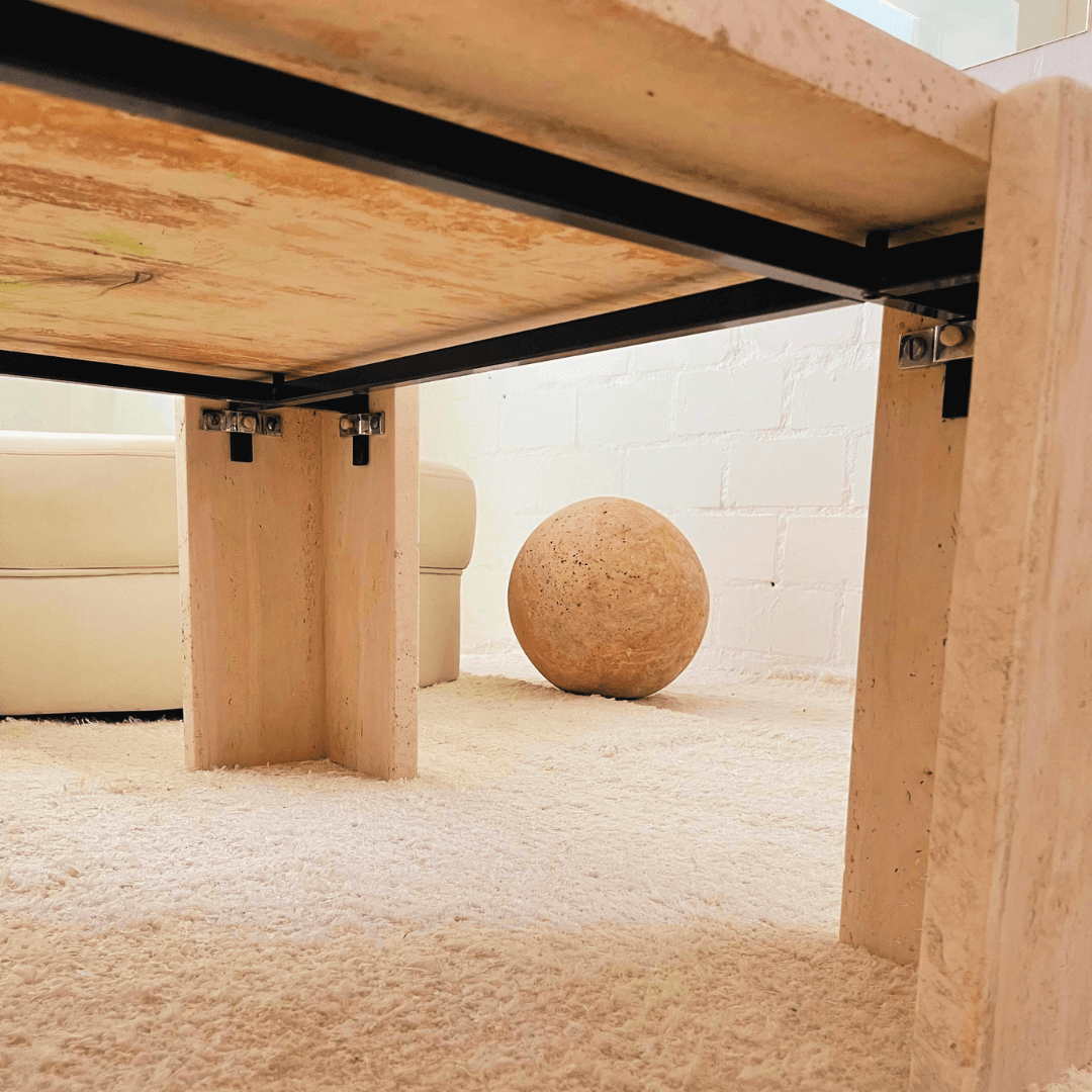 Under the Modern Rustic Travertine Coffee Table by Travertable, expert craftsmanship in leg supports is highlighted on a cream carpet. A large stone sphere sits near a white couch, with a white brick wall enhancing the rooms tranquility and natural beauty.