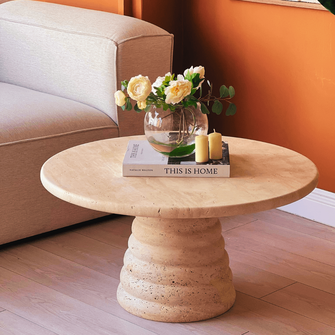 The Designer Travertine Marble Coffee Table by Travertable features a round beige top adorned with two yellow candles, a clear vase of white roses, and This Is Home. Its sculptural stone aesthetics complement the light beige sofa and pop against the orange wall, adding durability to any living space.