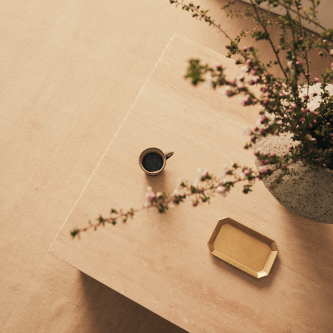A minimalist scene showcases a luxury home decor vibe with the Travertables Minimalist Travertine Accent Coffee Table. Accompanied by a small cup of black coffee, an elegant gold tray, and a vase of greenery and pink flowers, the serene setting exudes warmth and sophistication.