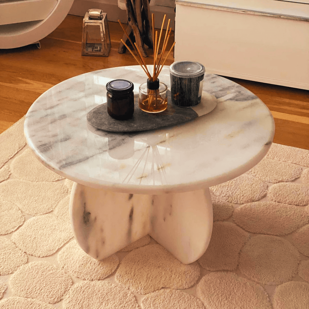 The Travertable Stone Essence Travertine Coffee Table, with its monolithic modern design, rests on a textured beige rug. Adorned with two dark candles and a reed diffuser set on a dark tray, the room highlights natural beauty with wooden flooring and minimal decor.