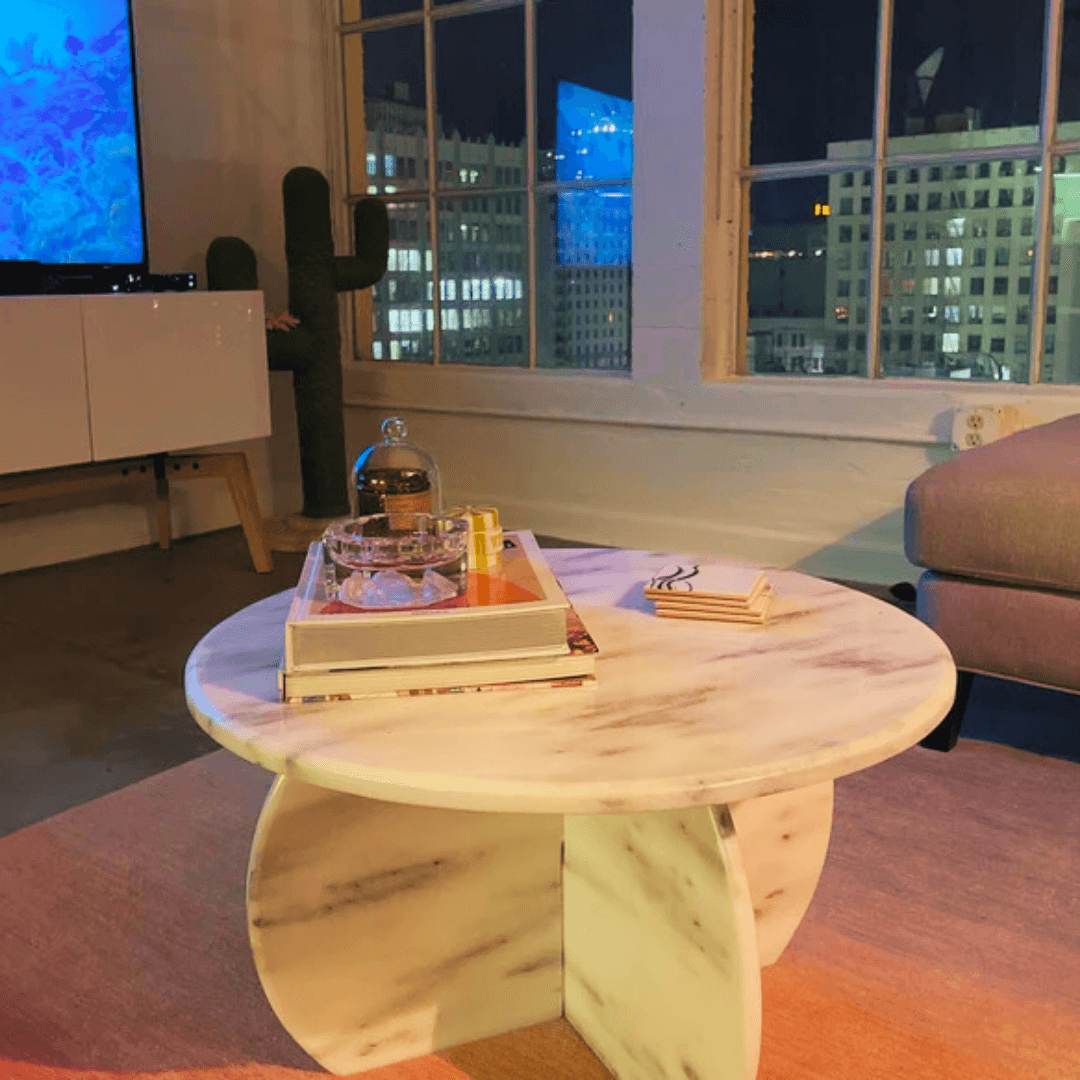 A cozy living room showcases a timeless design with a Travertable Stone Essence Travertine Coffee Table, styled with books and decor. Large windows display a vibrant cityscape, complemented by a cactus-shaped decor piece and TV, adding charm to the scene.