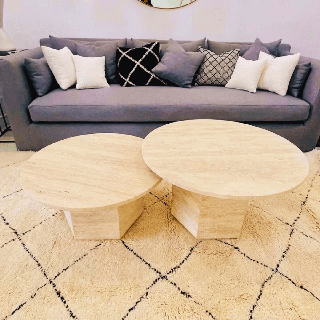 The Desert Serenity Travertine Coffee Table by Travertable, with its earthy, sculptural stone design and soft honed finish, complements a cream geometric-patterned rug. Behind it, a gray sofa featuring white and patterned throw pillows blends seamlessly with the light walls decorative accents.