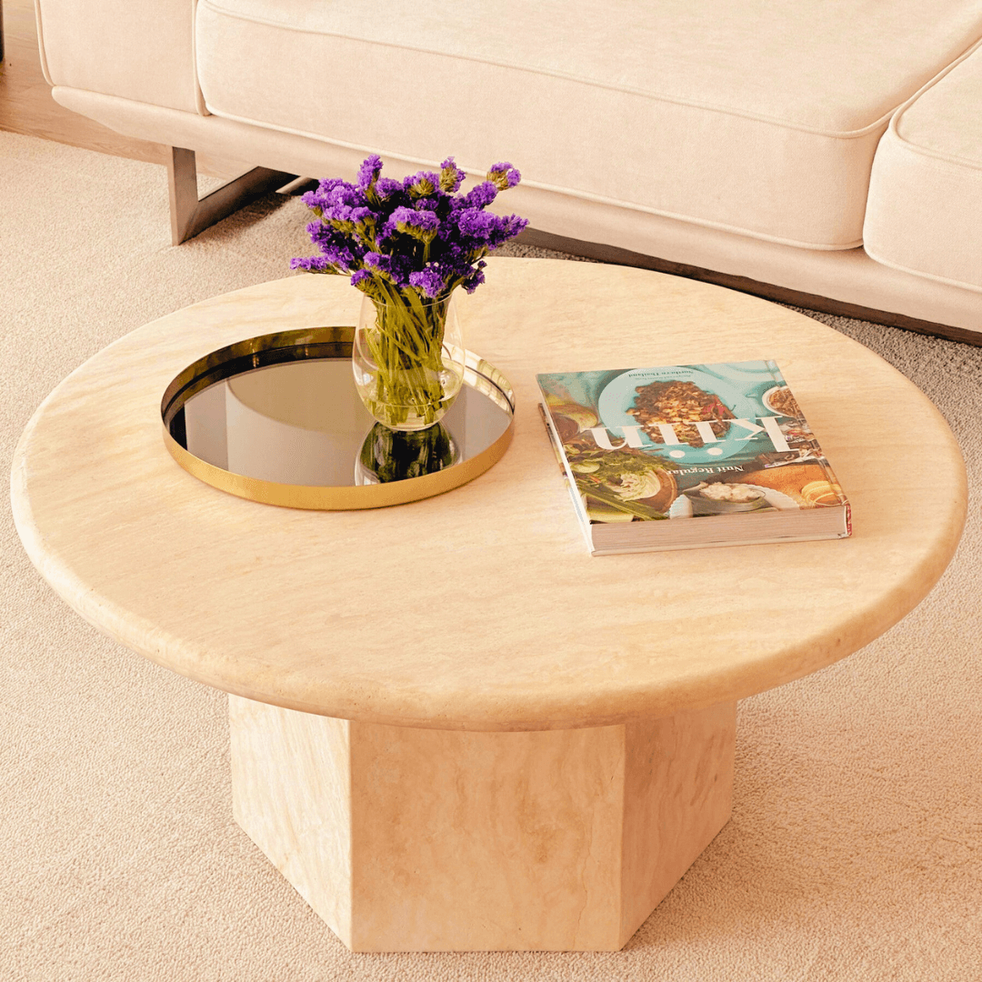 The Desert Serenity Travertine Coffee Table by Travertable, made from earthy stone with a honed finish, showcases a timeless design. It features a glass vase with purple flowers, a black and gold tray, and a colorful magazine on a light carpet in front of the cream sofa.
