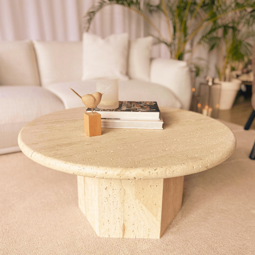 A Desert Serenity Travertine Coffee Table by Travertable, adorned with a small wooden bird sculpture and stacked books, graces the room. A white sofa and potted plants in the background enhance the timeless design, creating a cozy, modern living room atmosphere.