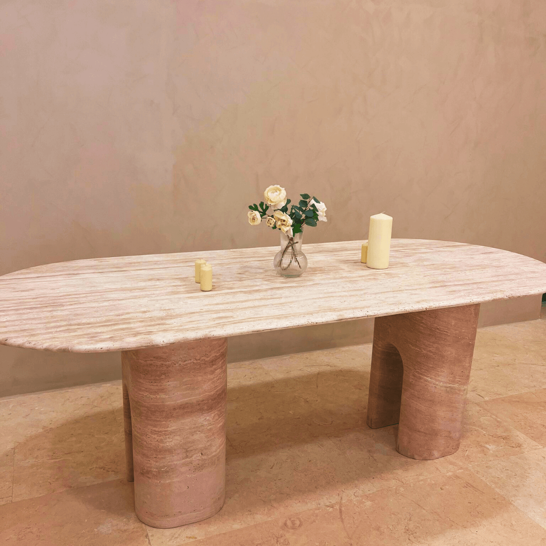 The Travertine Majesty Ellipse Table by Travertable, with its rounded legs and light hue, holds three cream candles and a glass vase of white flowers and green leaves, blending elegance with nature against a plain beige wall and light stone floor.