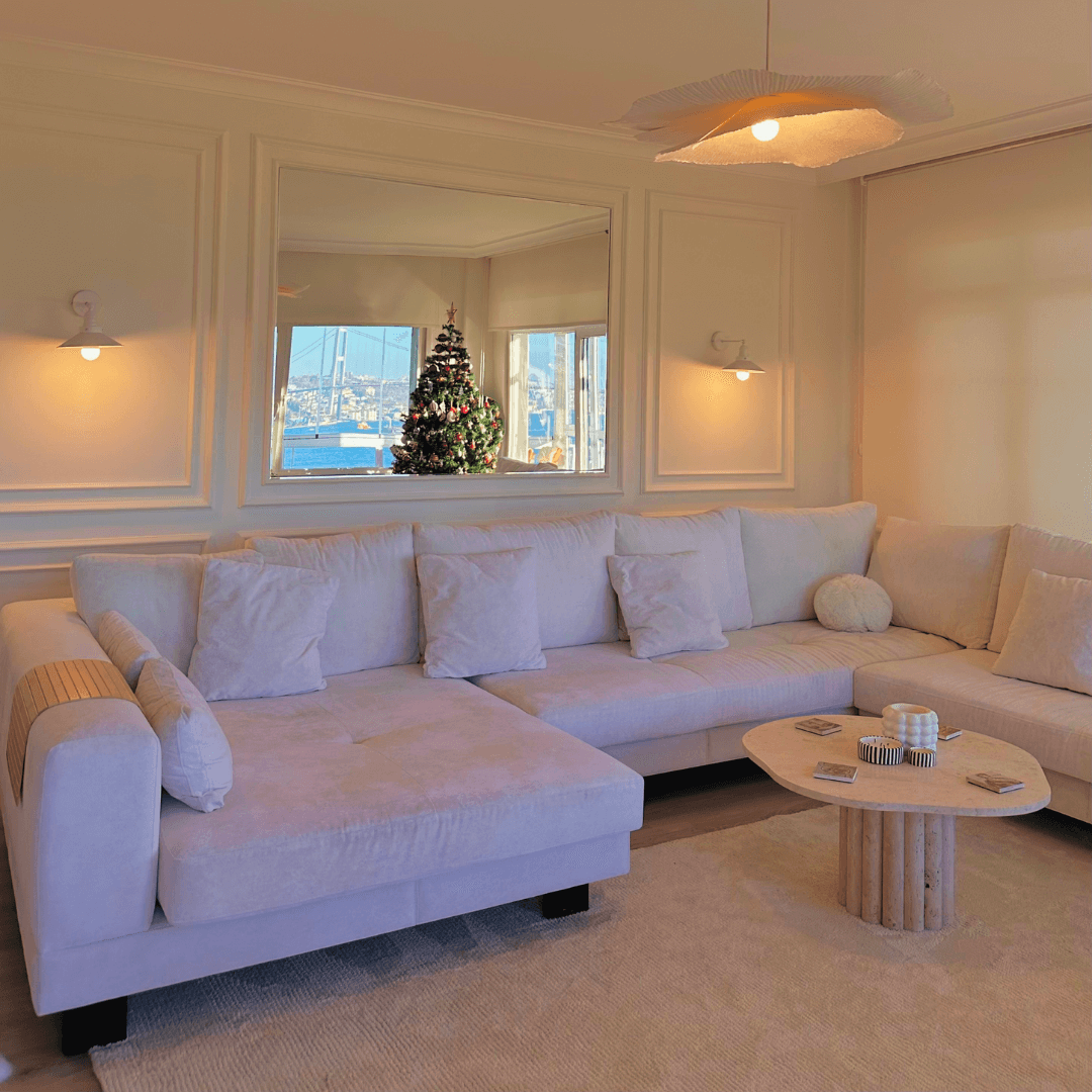 A cozy living room showcases a large beige sectional and a Timeless Stone Travertine Coffee Table by Travertable. A window reflects a decorated Christmas tree and marina view, while soft lighting from wall sconces enhances the warm ambiance.