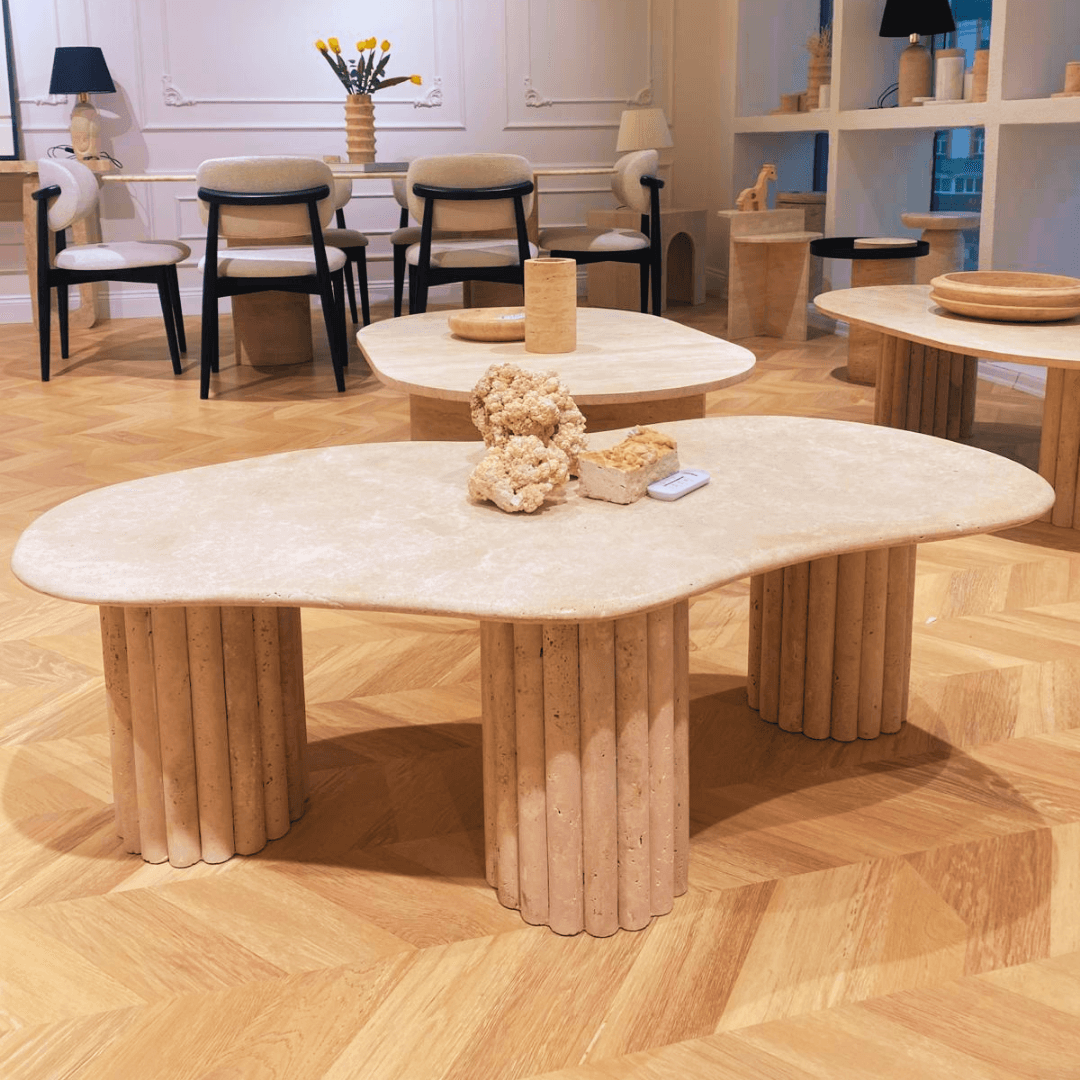 A stylish showroom with modern furniture showcases expert craftsmanship, featuring three beige stone tables with cylindrical bases. The prominent Terra Solace Travertine Coffee Table by Travertable displays decorative beige objects. A wood-paneled floor and well-lit interior add natural beauty amidst chairs and shelves.