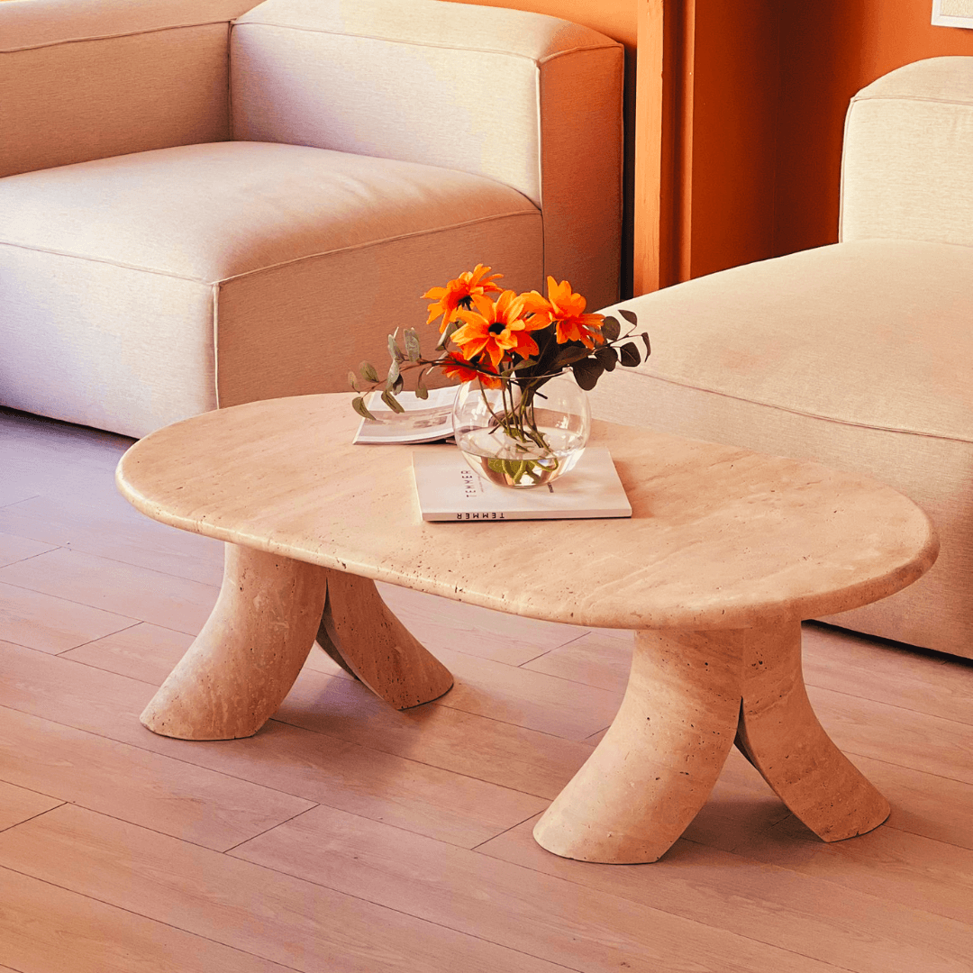 A minimalist living room features the Terra Elegance Travertine Coffee Table by Travertable as a luxury decor centerpiece with curved legs, topped with a glass vase of orange flowers and two white books. Beige armchairs and a wooden floor create a warm, cozy atmosphere.