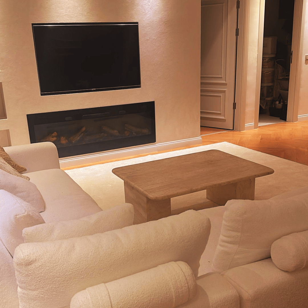 A cozy living room with a plush white sofa facing a flat-screen TV above a modern fireplace features Travertables Limestone Luxury Coffee Table, showcasing timeless natural beauty and sculptural design. Warm lighting and a partially open door reveal an elegant connection to another room.