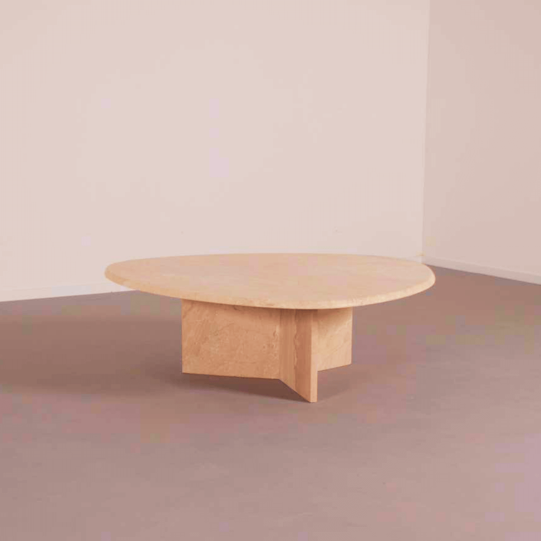The Cavern Charm Travertine Coffee Table by Travertable, a vintage-inspired oval piece with raw appeal, features a sturdy base complementing the smooth travertine stone top. It rests on a gray floor, beautifully set against a plain light-colored wall.
