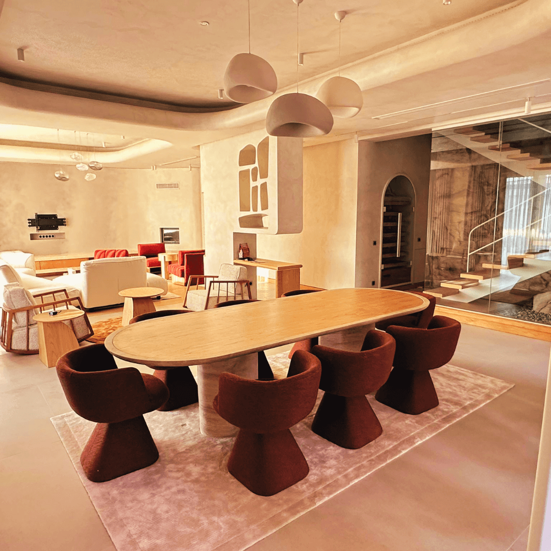 A modern dining area showcases The Harmony Travertine and Walnut Dining Table by Travertable, surrounded by red chairs on a soft rug. It features a white wavy ceiling, contemporary pendant lights, cozy sofas, and a visible staircase that enhances its customizable design.