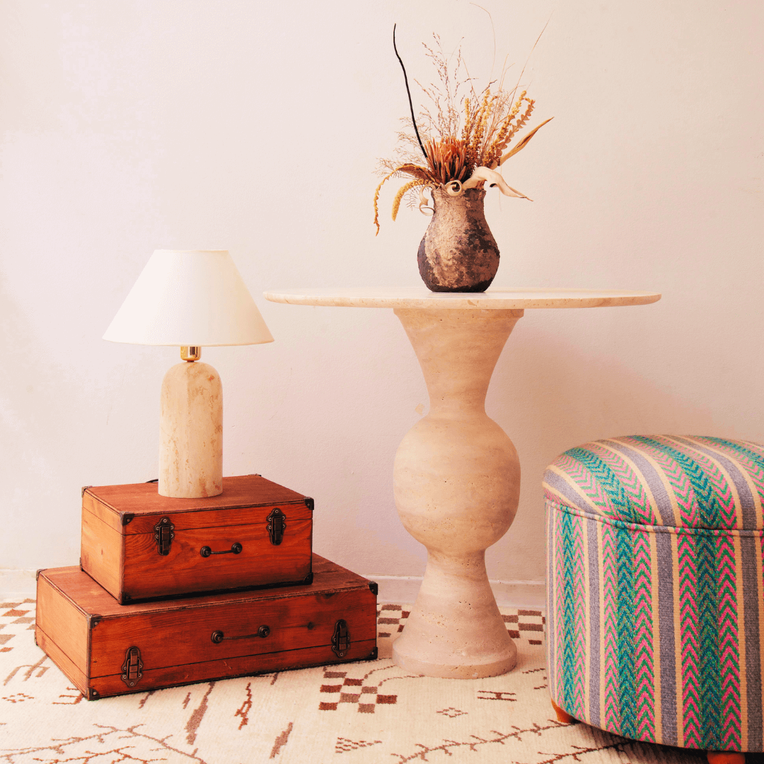 Beside two stacked wooden boxes topped with a lamp, The Hourglass Round Travertine Dining Table by Travertable holds a vase of dried flowers. Nearby is an upholstered stool with colorful zigzag patterns on a patterned rug. The plain white wall enhances the minimalist decor and highlights the tables hourglass base design.