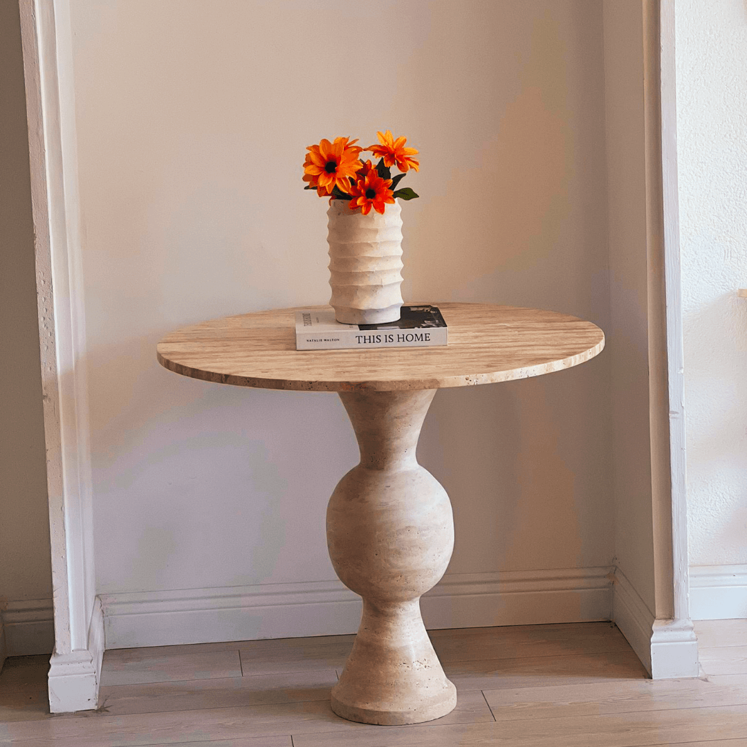 A minimalist setting features Travertables The Hourglass Round Travertine Dining Table against a neutral-hued wall, creating a serene vibe. It holds a white vase with bright orange flowers and two books, including This is Home.