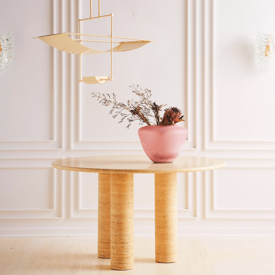 The Trio Round Travertine Dining Table by Travertable with dual cylindrical legs is set against white paneled walls. A pink vase with dried flowers sits atop, while a modern geometric pendant light illuminates the light wood floor below.