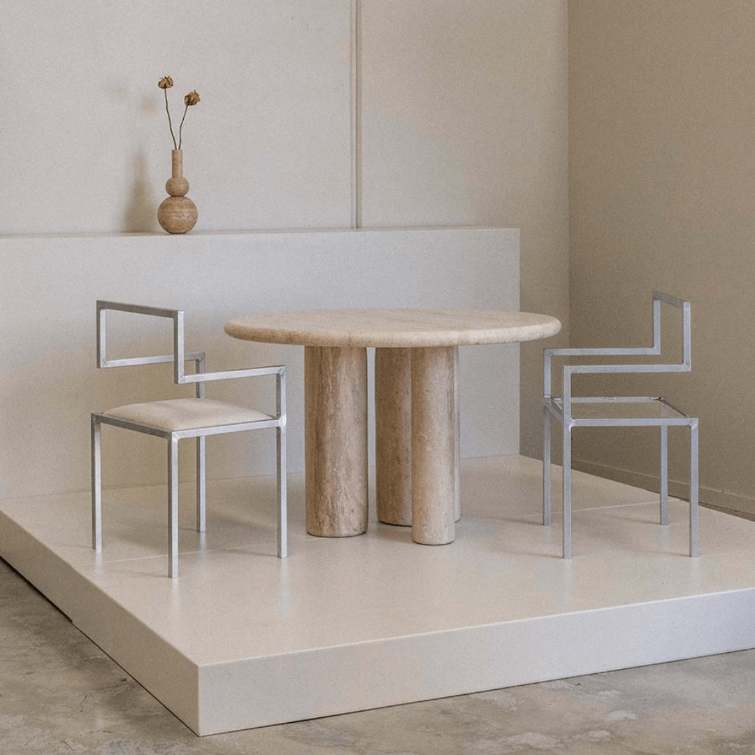 A minimalist space showcases The Trio Round Travertine Dining Table by Travertable, accompanied by two geometric metal chairs on a raised platform. A small vase with dry plants graces the table, amplifying the neutral and modern aesthetics.
