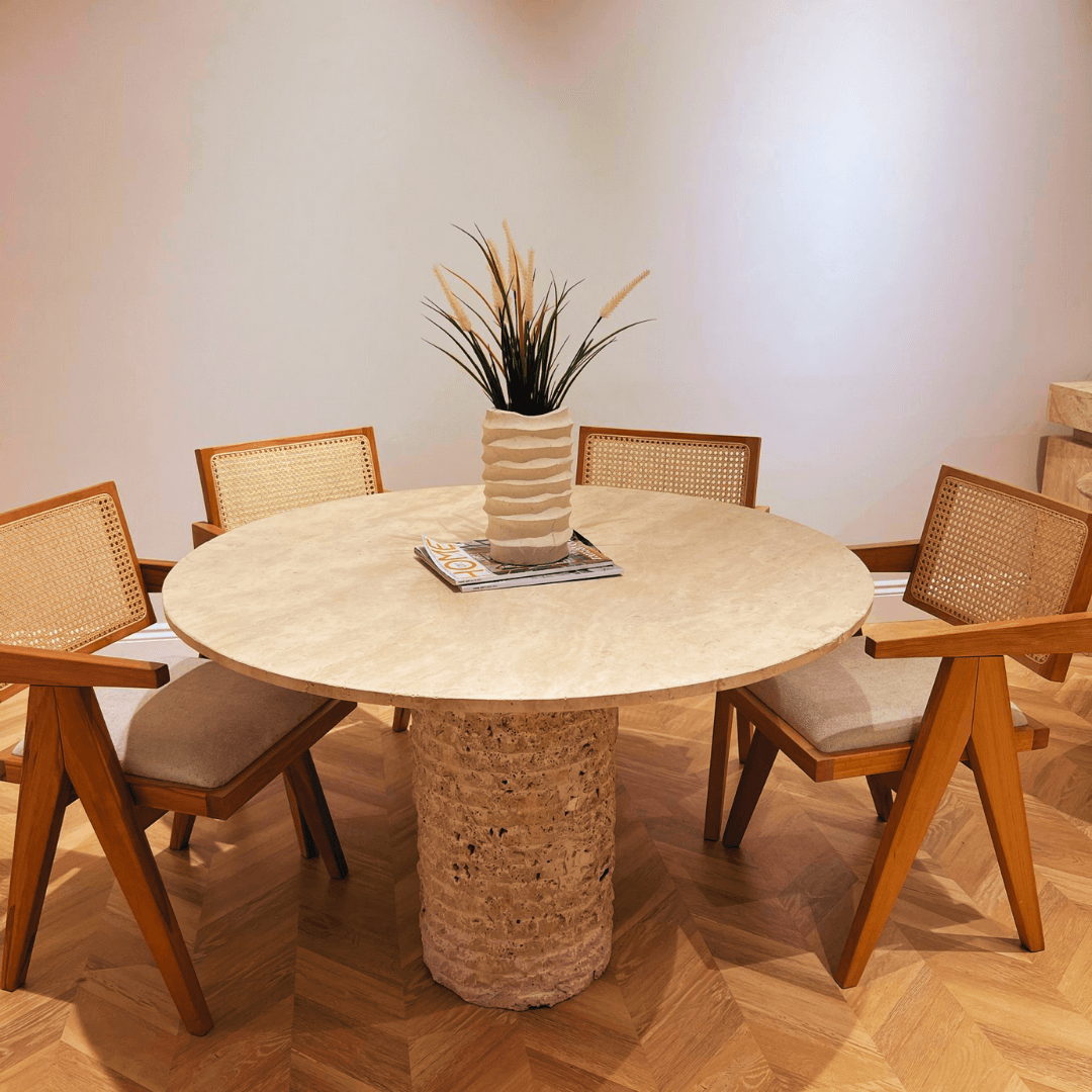 The Rustic Round Travertine Dining Table by Travertable features a textured pedestal stone base on wooden parquet. Four wooden chairs with woven backs and cushions surround the customizable tabletop, decorated with a plant and magazines.